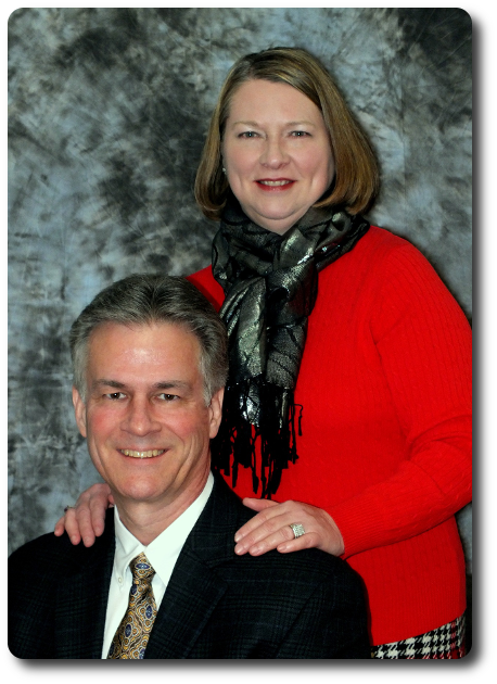 Pastor Bill Pickel seated in a suit, with Mrs. Pickel standing beside him in a red sweater